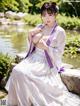 A woman in a white and purple kimono sitting on a rock by a pond.